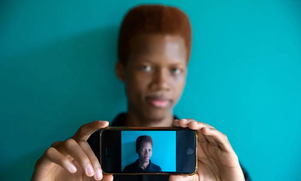 A person taking a live image of themselves with their phone. Their face can be seen behind the phone and also in the front screen of the phone taking a photograph.