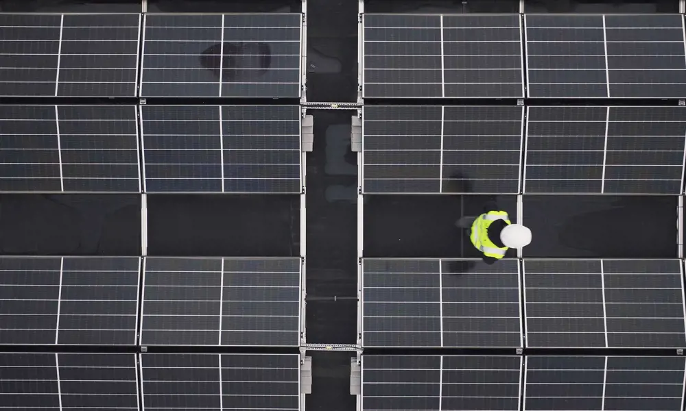An aerial view of solar panels on a roof with an apprentice engineer in a high visibility jacket and helmet inspecting them.