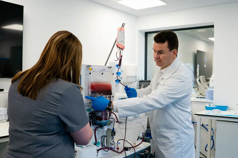 A man wearing a white coat and blue gloves stand in fron of a machine directing a person with long brown hair who has their back to the camera