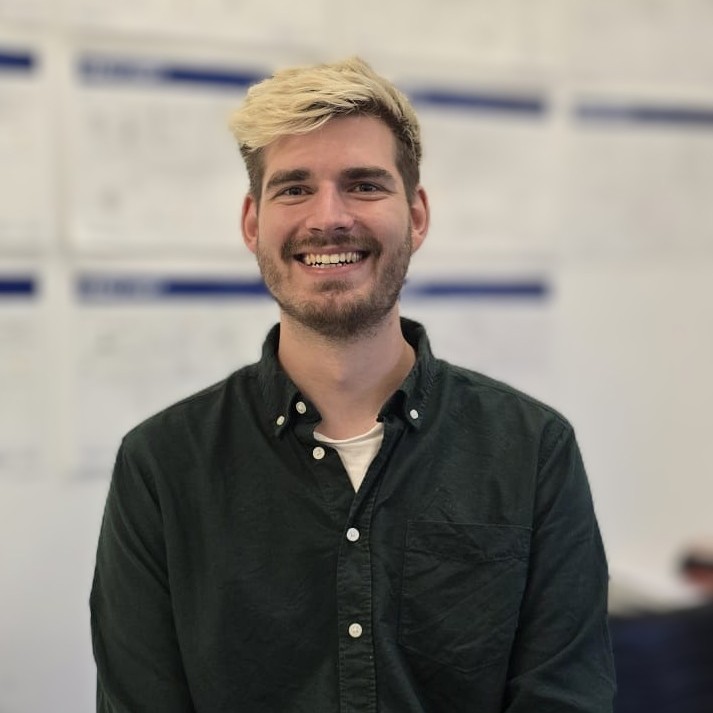 A person with light blond hair wearing a dark green button‑up shirt sits indoors in front of a wall covered with charts and diagrams.