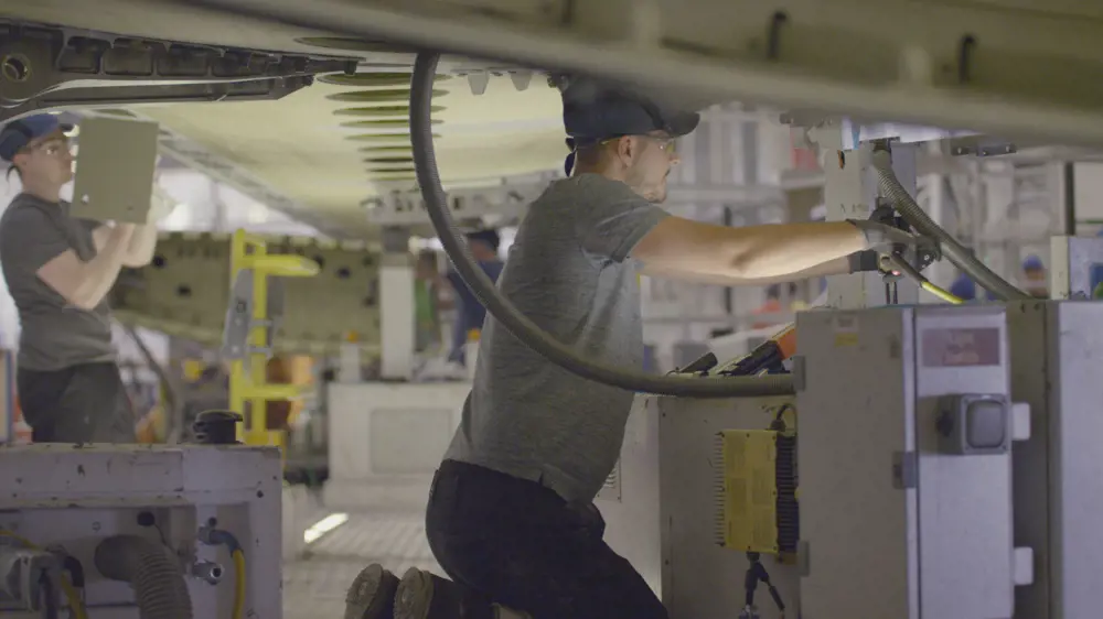 An aircraft fitter engineer kneeling, working inside a factory under an aircraft wing.