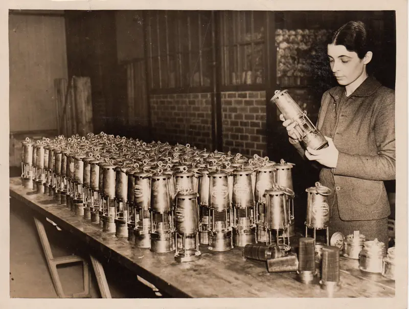 A monochrome photograph of Monica Maurice holding a lamp on a table that has rows of lamps on top of it that have been produced by Wolf Safety Lamp Company. 