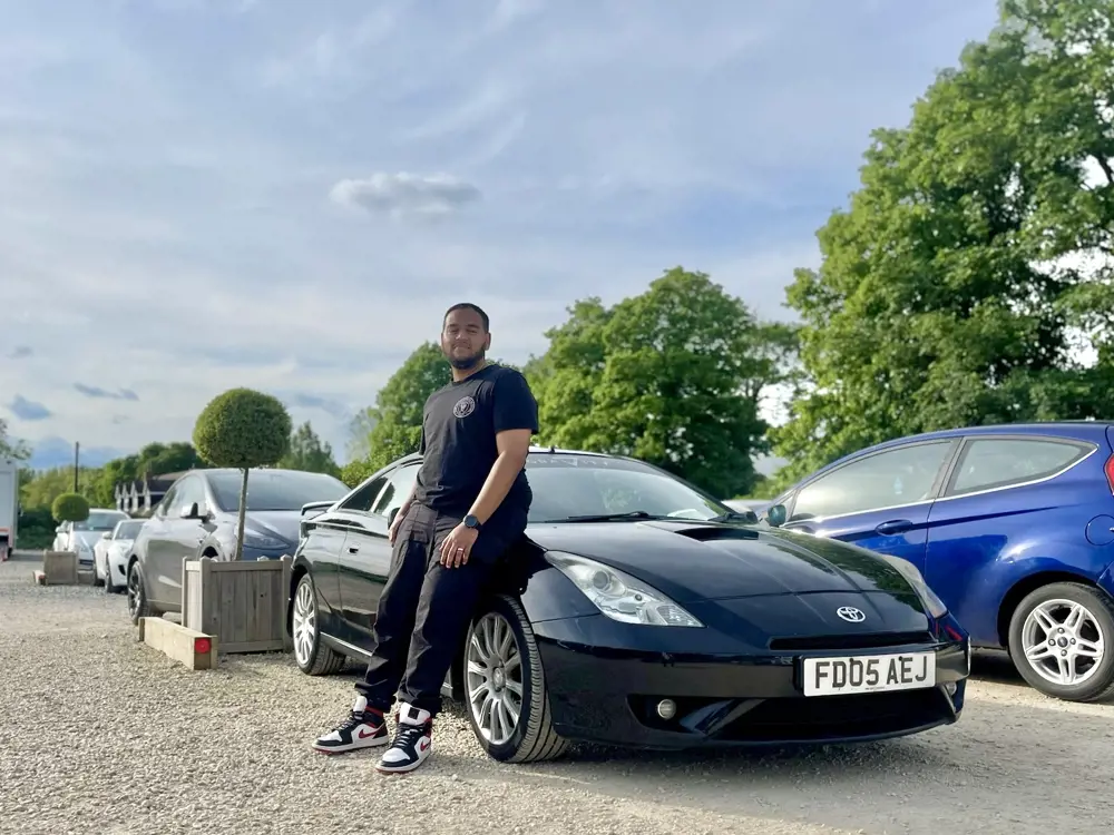 A man smiles, leaning against a Toyota car in a car park on a sunny day