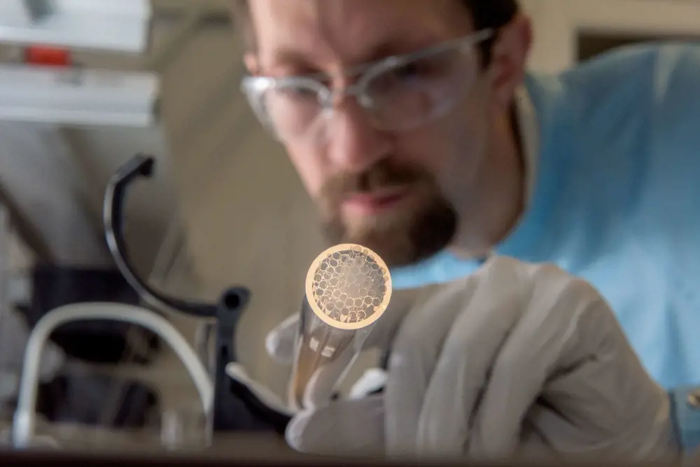 A researcher in a lab holding a stack jacket tube which has a glowing outer diameter containing many smaller hollow capillary tubes that is being pointed at the camera.
