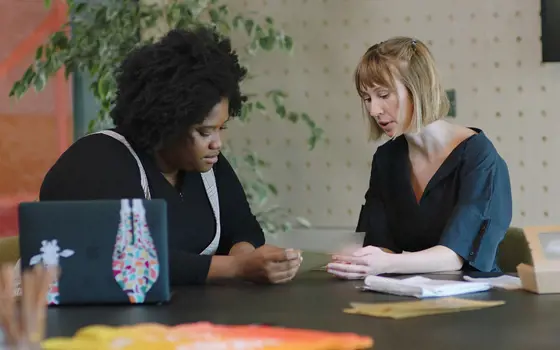 A Black woman and a white woman sit opposite each other at a table, looking down at a thin piece of transparent material they are both holding