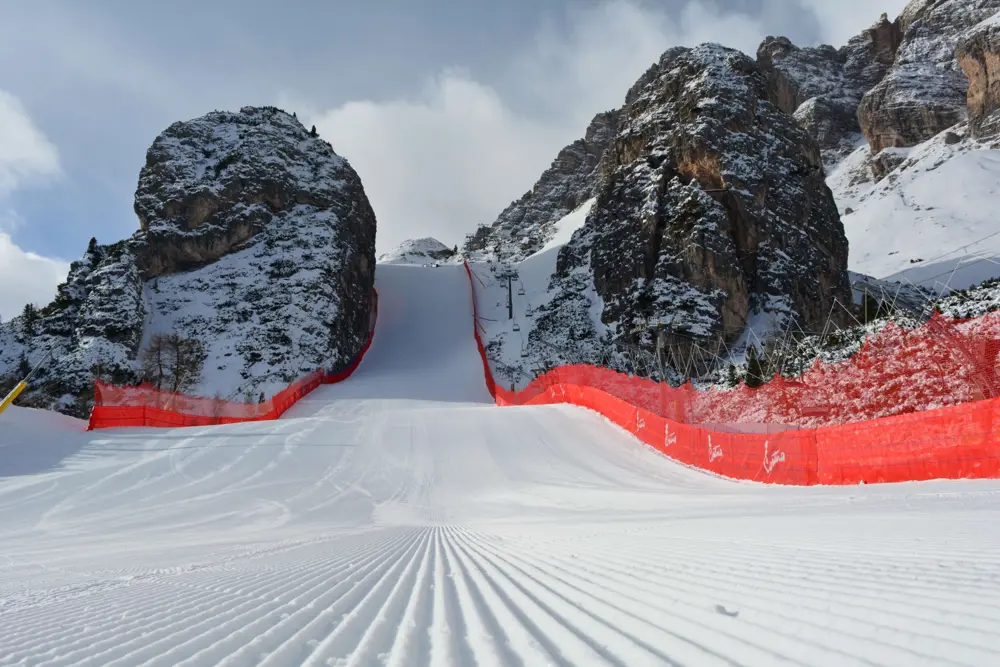 A freshly groomed ski slope runs steeply between two tall rocky cliffs, bordered by bright red safety fencing.