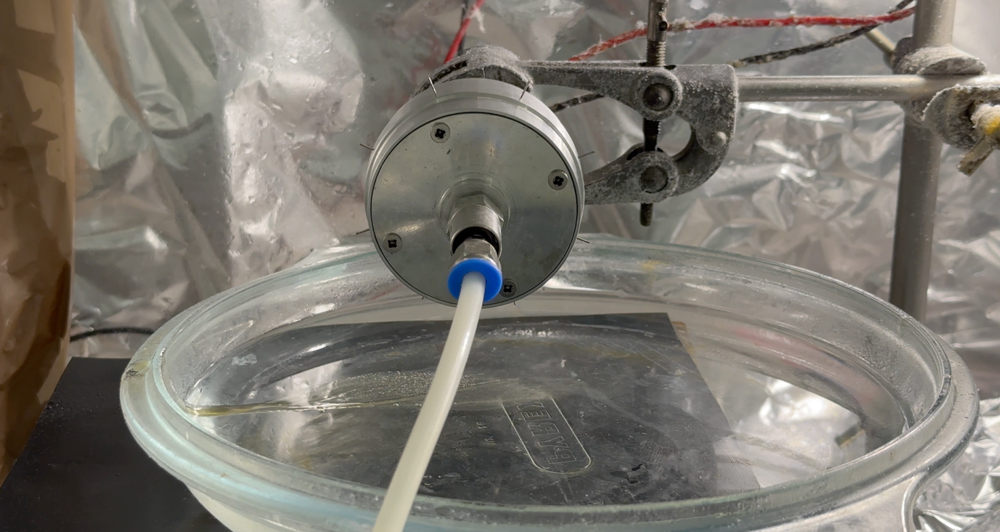 Close-up of a laboratory setup showing a metal spinning head connected to tubing positioned above a glass container, with foil and structural supports surrounding the apparatus.