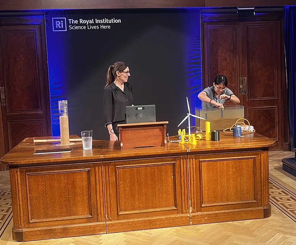 A woman wearing a dark shirt and glasses, stands behind a large wooden desk, upon which there is a jug of water, laptop and miniature wind turbine. She is looking at a second woman with dark hair who is bending over to look into a tank of water 