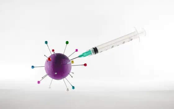 A syringe with the needle sticking into a purple sphere with long pins sticking out of it, which is a model of a covid virus particle