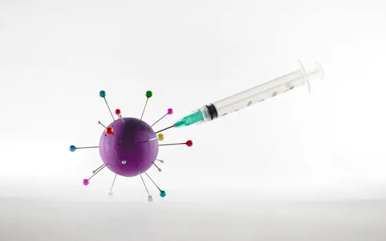 A syringe with the needle sticking into a purple sphere with long pins sticking out of it, which is a model of a covid virus particle