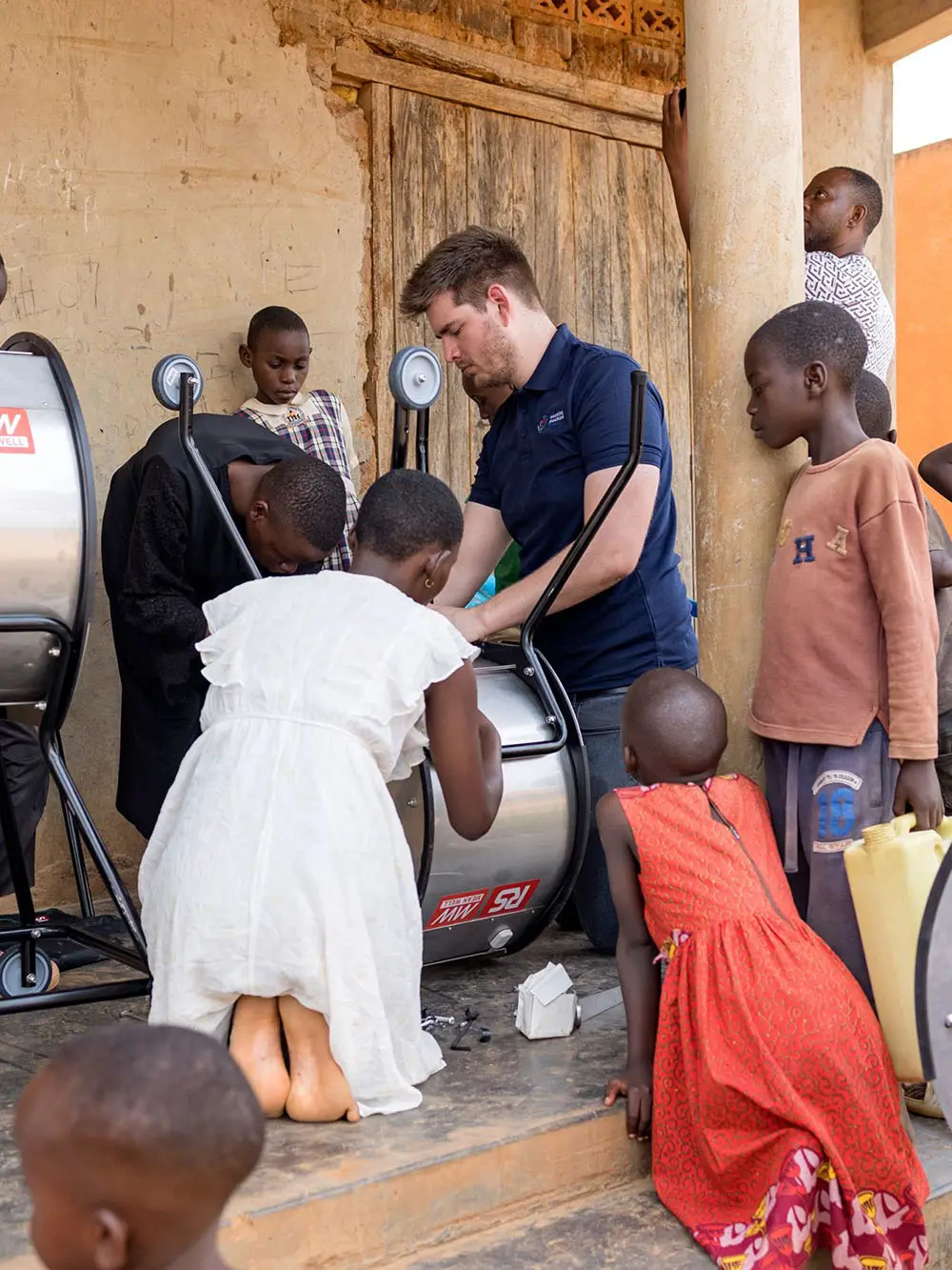 A group of children and an adult gather around a manual washing machine outside a building, watching as someone demonstrates how to use it.