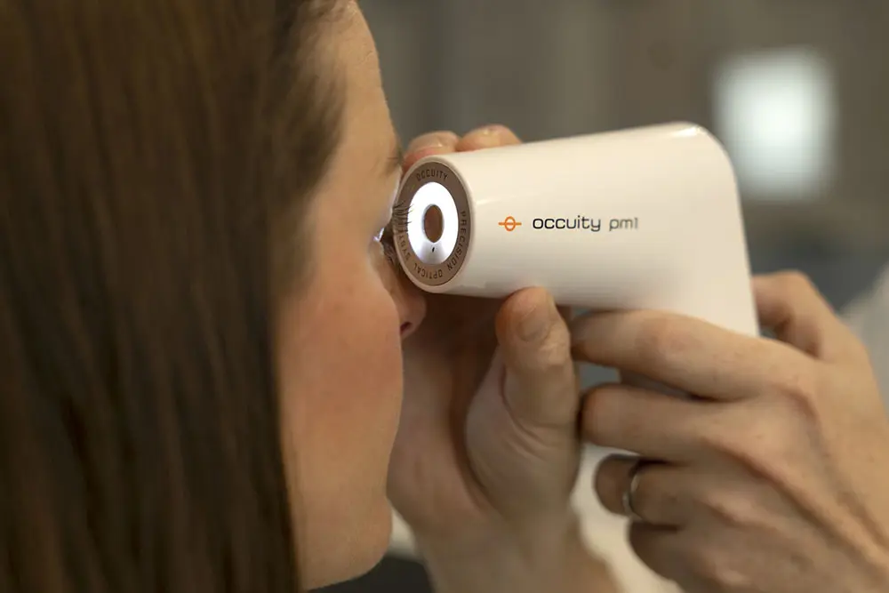 Close-up of a handheld white medical device labelled “Occuity PM1” being positioned near the side of a person’s head, likely for an eye health or diagnostic scan