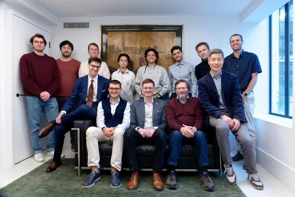 A group of people posed together in a bright meeting room, with some seated on a sofa and others standing behind, in front of a large framed artwork on the wall.