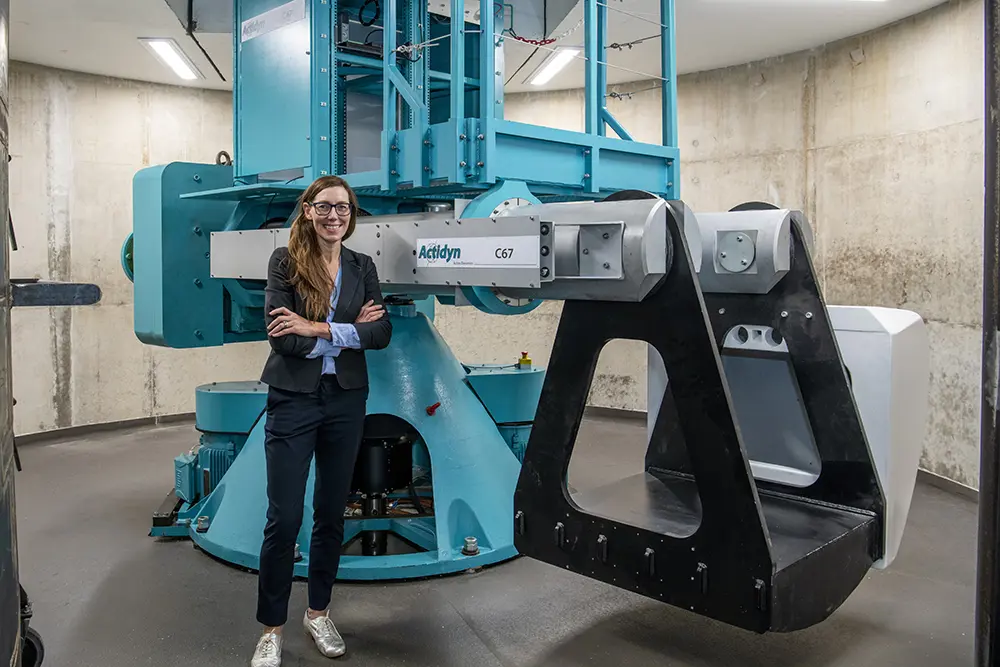 A woman with long dark hair wearing glasses, a suit and silver shoes stands in front of a large blue centrifuge in an industrial setting