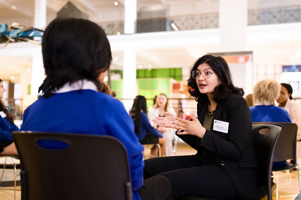 A person leads a small group discussion with students seated in a museum atrium, with other groups and exhibits visible in the background.