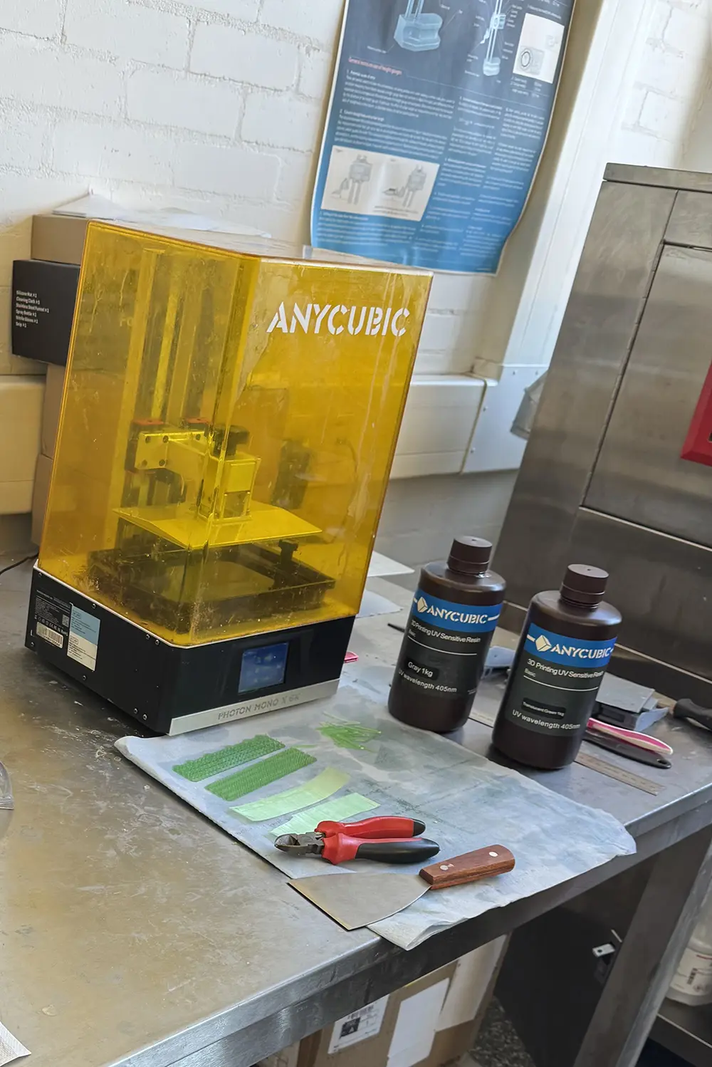 A 3D printer sits on a worktop in an industrial setting alongside two black bottles of resin, and some green strips, a pair of pliers and a spatula on a piece of fabric