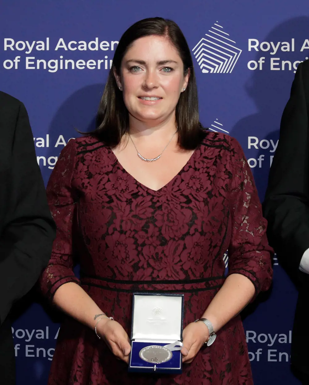 A women with brown hair, wearing a maroon formal dress stands in front of a blue background holding a silver medal in a presentation box