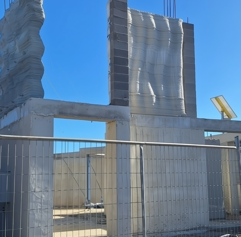 3D‑printed concrete walls of a multi‑storey house under construction, viewed behind a safety fence.