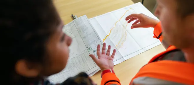 Close-up of two individuals reviewing a detailed engineering plan on a table, with one wearing a high-visibility jacket and gesturing toward the design, illustrating collaboration and forward-looking infrastructure planning.