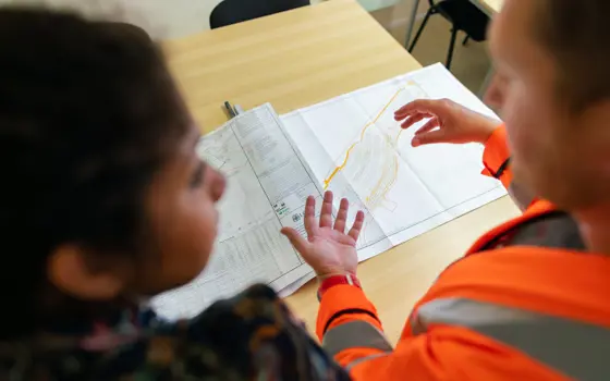 Close-up of two individuals reviewing a detailed engineering plan on a table, with one wearing a high-visibility jacket and gesturing toward the design, illustrating collaboration and forward-looking infrastructure planning.