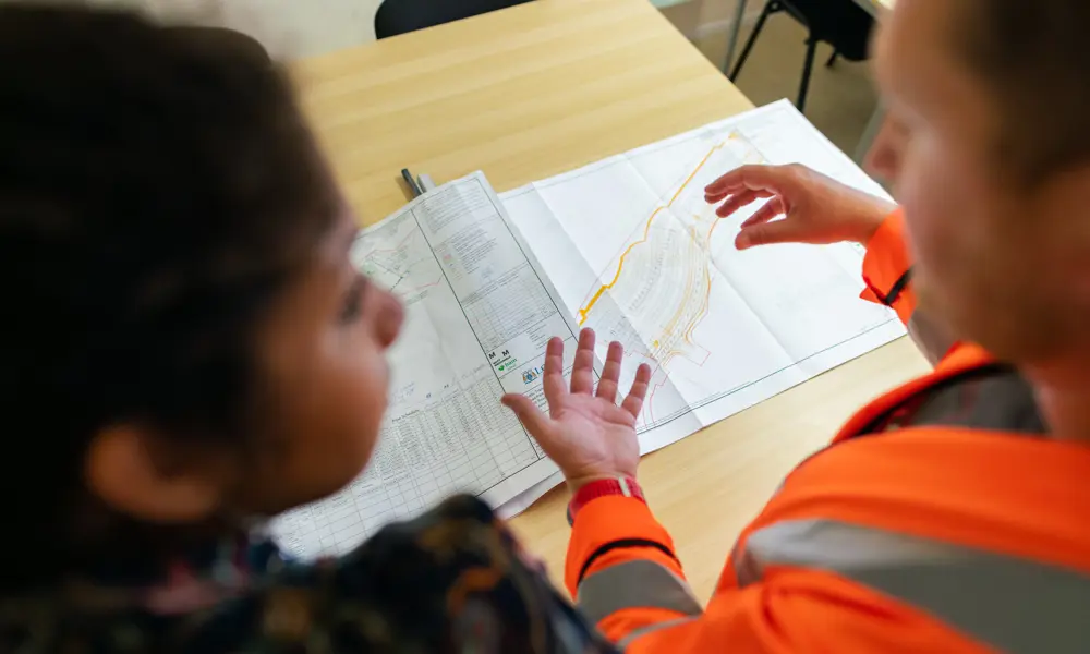 Close-up of two individuals reviewing a detailed engineering plan on a table, with one wearing a high-visibility jacket and gesturing toward the design, illustrating collaboration and forward-looking infrastructure planning.