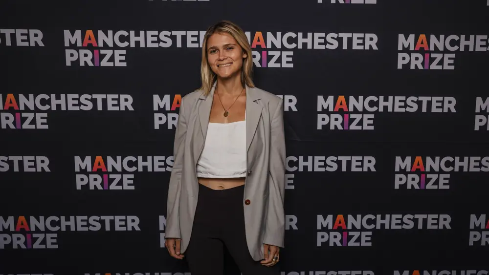 A blonde woman smiles for a photo in front of a backdrop reading "Manchester Prize"