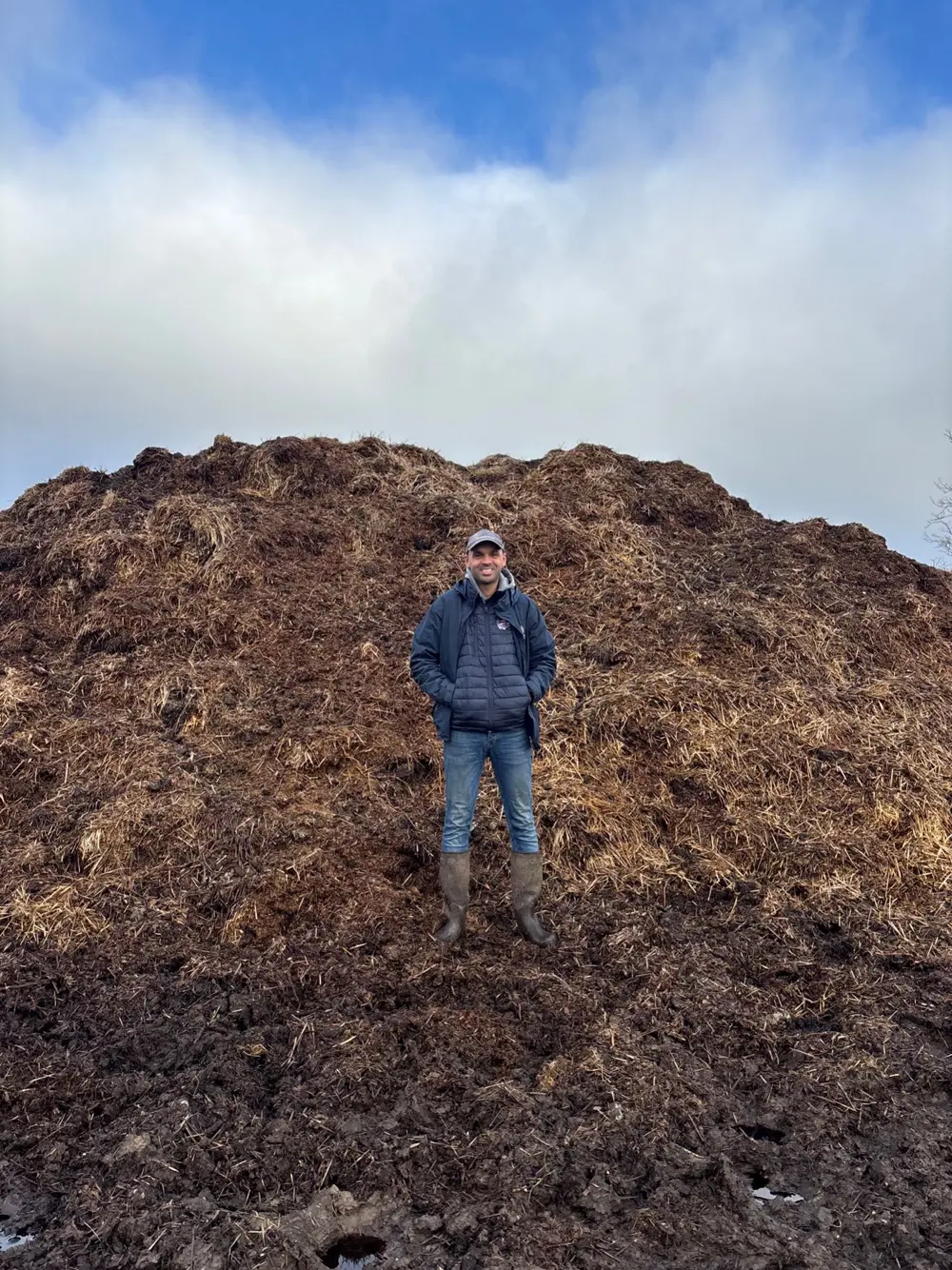 Perry Robinson wearing outdoor clothing and wellies standing in front of a large mound of brown manure, under a cloudy sky.