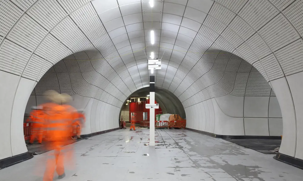 The inside of the underground Elizabeth Line station while it was under construction with glass fibre reinforced concrete cladding panels lining the walls. A blurry worker in construction gear is walking through the tunnel. 