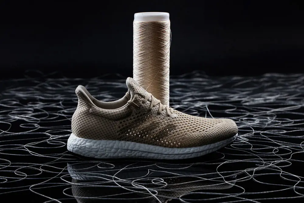 Adidas trainers that are attached to a roll of thread, resting on a floor containing pieces of coiled thread.