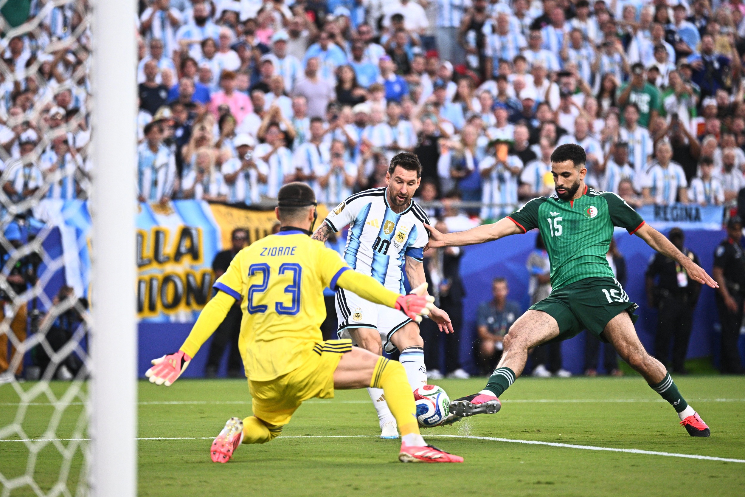 Lionel Messi of Argentina shoots the ball past Rayan Aït-Nouri (Algeria) and goalkeeper Luca Zidane (Algeria) to score, in front of a packed crowd in blue and white stripes.