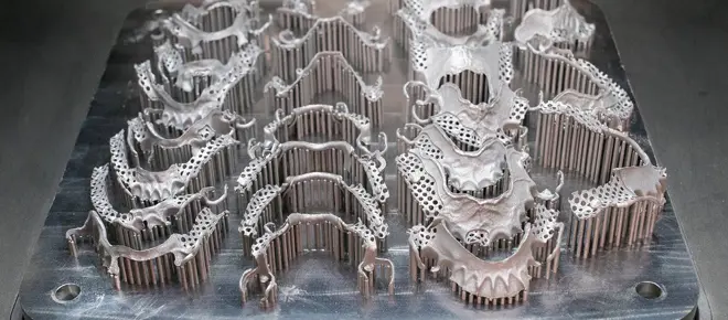 A flat build plate containing multiple metal dental products attached to the plate by many vertical pins.