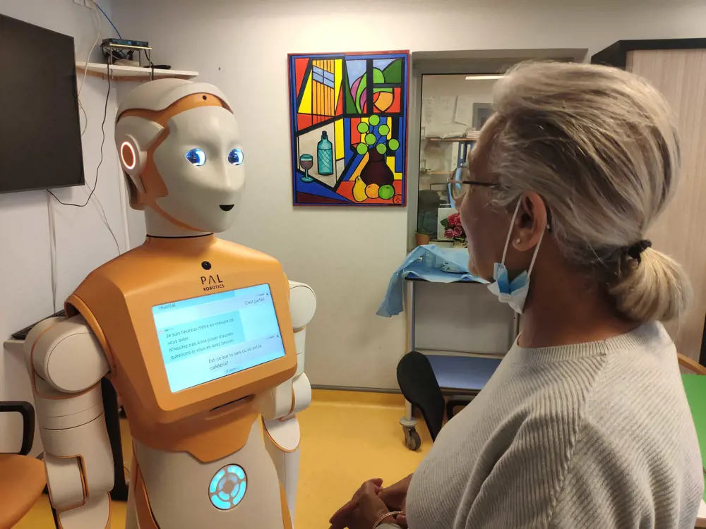 An older woman speaks to a robot with an orange torso and white head, body and arms