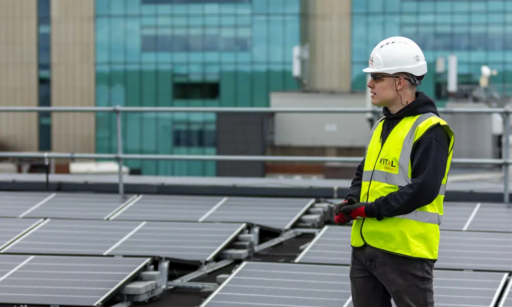 Harvey Hudson wearing a hard hat and yellow jacket from Vital Energi, standing on the roof of the Christie Hospital which is covered in solar panels.