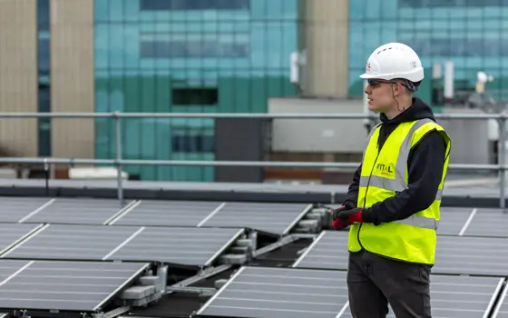 Harvey Hudson wearing a hard hat and yellow jacket from Vital Energi, standing on the roof of the Christie Hospital which is covered in solar panels.