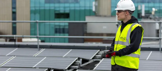 Harvey Hudson wearing a hard hat and yellow jacket from Vital Energi, standing on the roof of the Christie Hospital which is covered in solar panels.