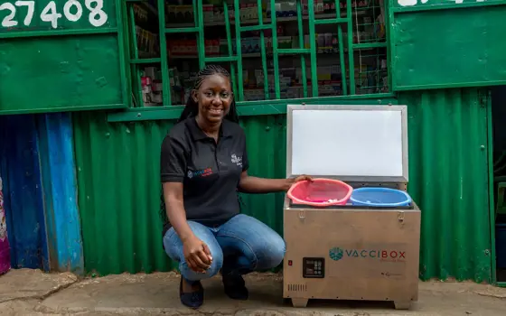 A woman crouches, smiling, next to a portable metal fridge with "Vaccibox" written on the front.