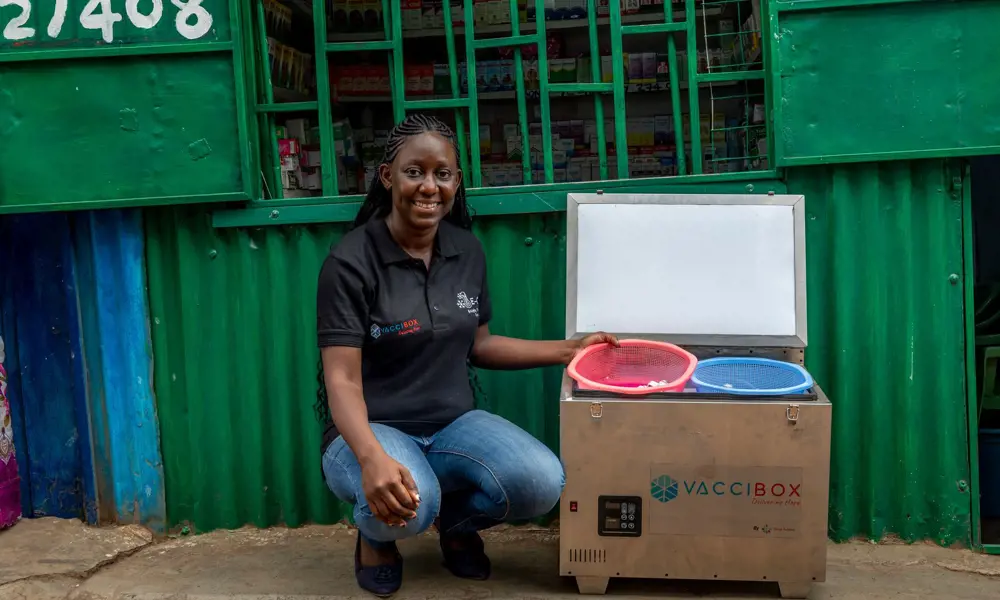 A woman crouches, smiling, next to a portable metal fridge with "Vaccibox" written on the front.