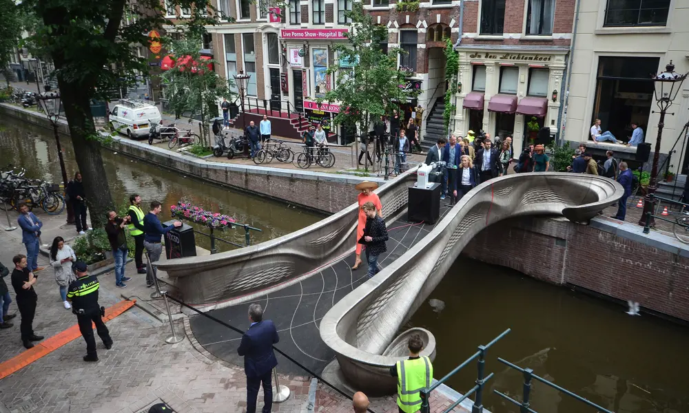 A curvy metal bridge across a canal in Amsterdam is seen from above. A group of people are walking across it, led by Queen Maxima of the Netherlands.
