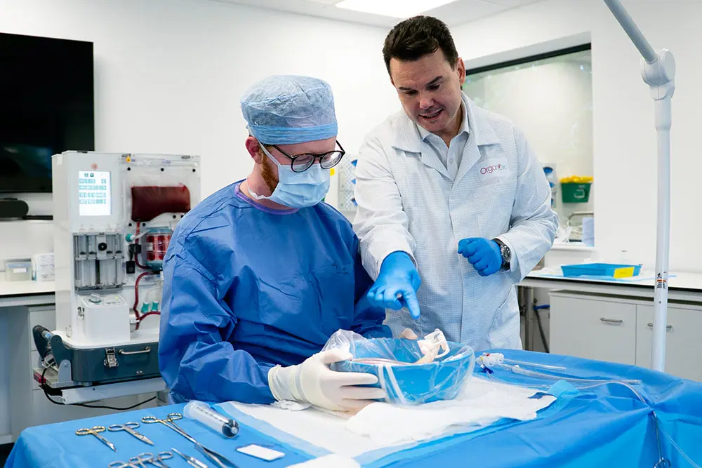 Two men in a clinical area, one wearing blue scrubs including a hair net and a face covering and one wearing a white jacket with blue gloves, look down into a blue bowl