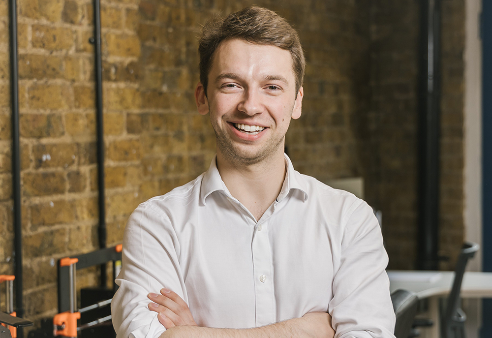 Portrait of a person in a white shirt standing indoors with arms crossed, against an exposed brick wall background.