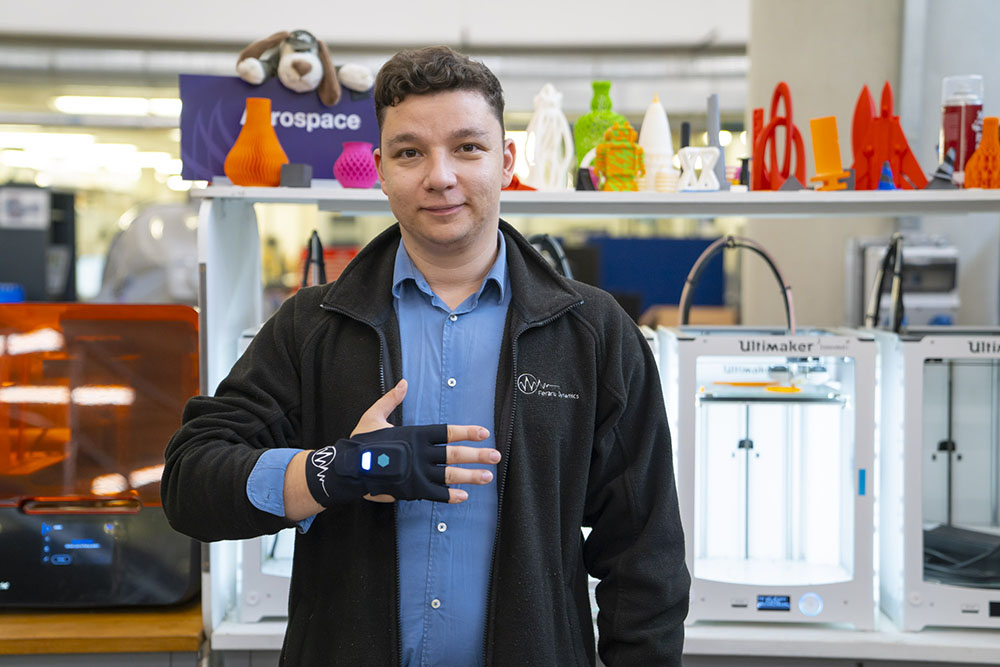 A person in a workshop demonstrates a high‑tech glove device, standing in front of shelves filled with 3D‑printed models and 3D printers.