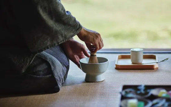 Hands whisking tea in a ceramic bowl during a traditional Japanese tea ceremony, with utensils on a tatami mat.