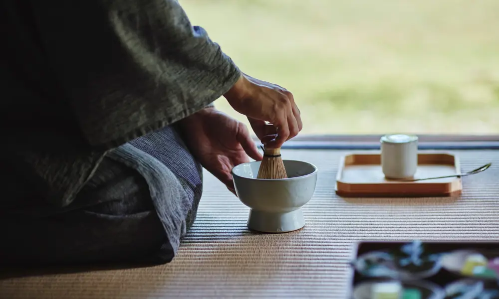 Hands whisking tea in a ceramic bowl during a traditional Japanese tea ceremony, with utensils on a tatami mat.