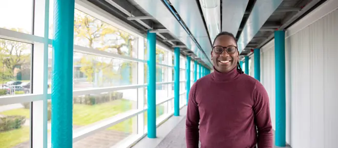 James Dornor wearing glasses and a burgundy jumper stands smiling in a bright corridor with turquoise columns and large windows.