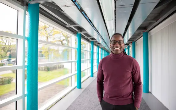 James Dornor wearing glasses and a burgundy jumper stands smiling in a bright corridor with turquoise columns and large windows.