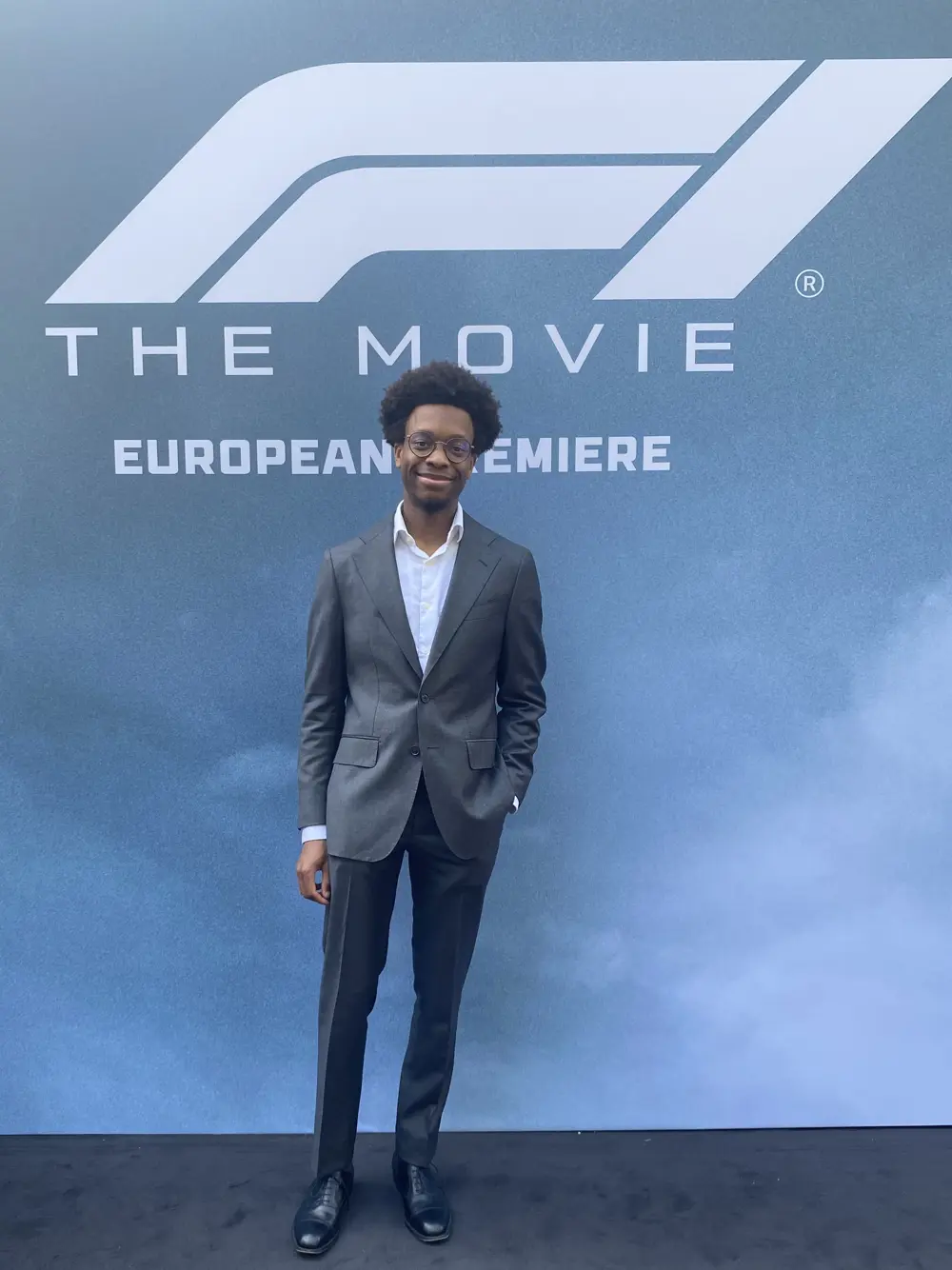 A man in a blue-grey suit posing for a photo in front of a backdrop that reads 'F1 The Movie, European Premiere'.