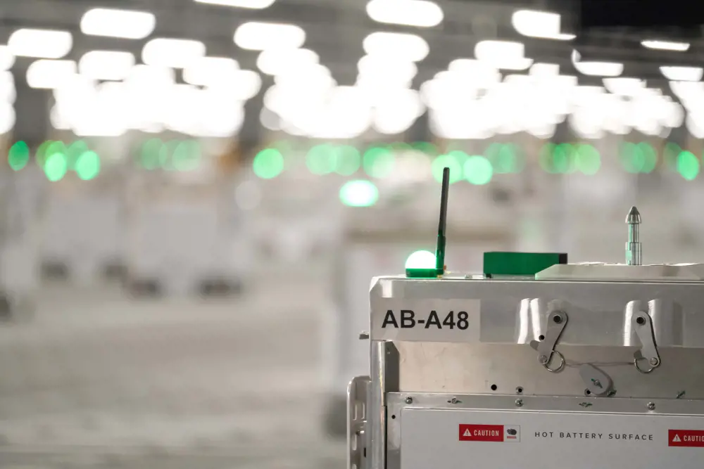 A close-up of a cube-shaped robot with a green light on it. Other robots on wheels can be seen out of focus in the background of Erith warehouse. 