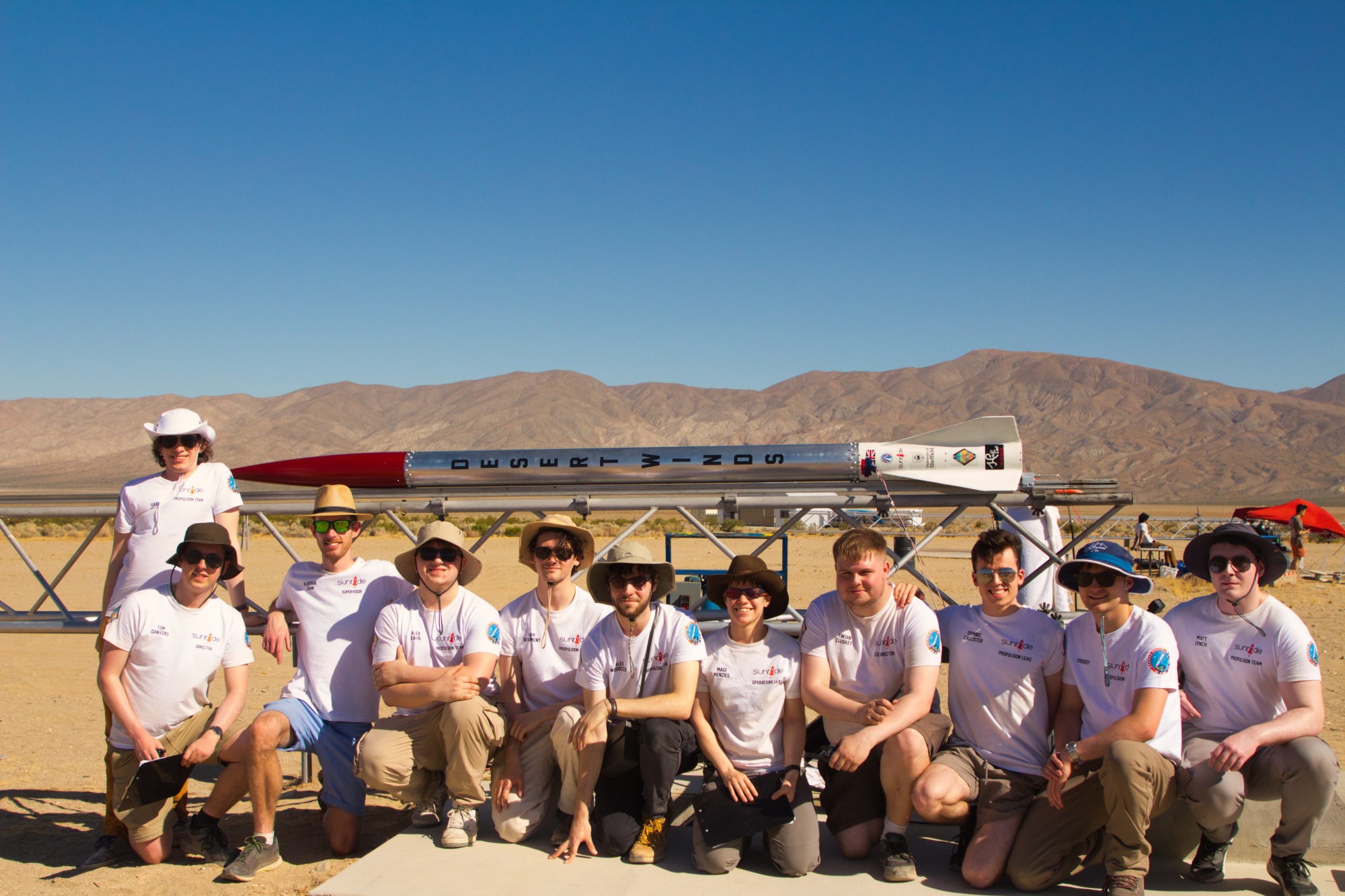 A group of people standing and kneeling in front of a rocket on a launch frame in a desert, with mountains in the background.