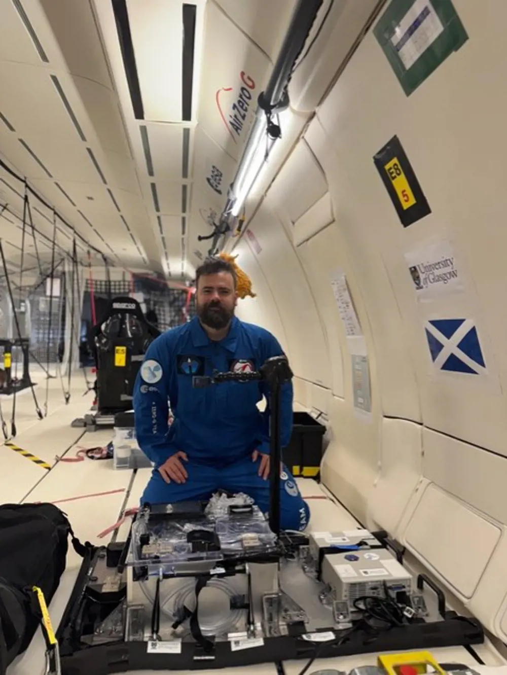 A bearded man in a blue boiler suit, kneeling in front of an experimental setup tethered to the floor.