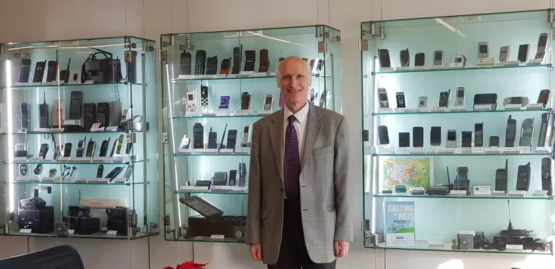Stephen Temple standing in front of a cabinet case with the mobile phone collection that he created at the University of Surrey,